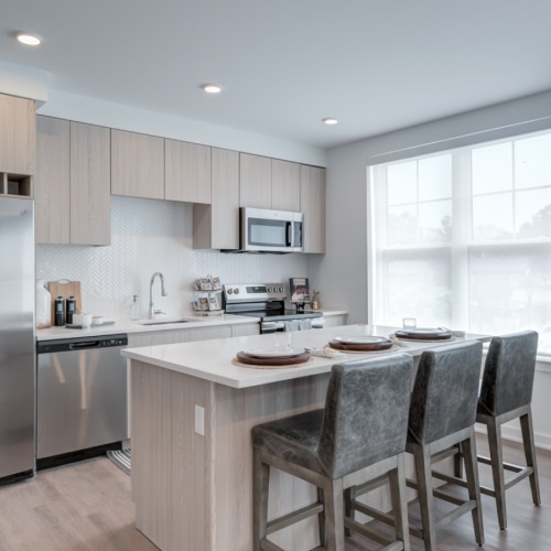 Modern apartment kitchen with island, bar stools, stainless steel appliances, and light wood cabinetry